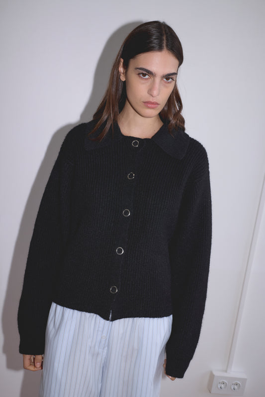 A woman with straight brown hair wears the Stud Cardi Knit - Black in an oversized fit and light blue striped pants, standing against a plain white wall with a slightly serious expression.