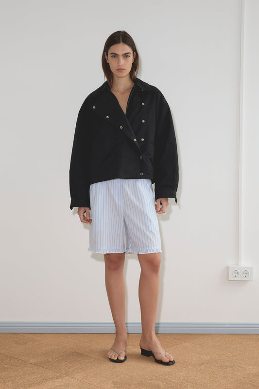 A model wearing the Lapel Jacket - Black with silver snap buttons, light blue pinstripe bermuda shorts with frayed hem, and black chain-detail sandals stands against a white wall.