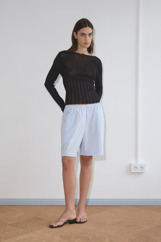 A woman stands indoors against a white wall, wearing the Deiji Studios Open Work Longsleeve in black, light blue striped knee-length shorts, and black sandals. Her hands are behind her back and she maintains a neutral expression.