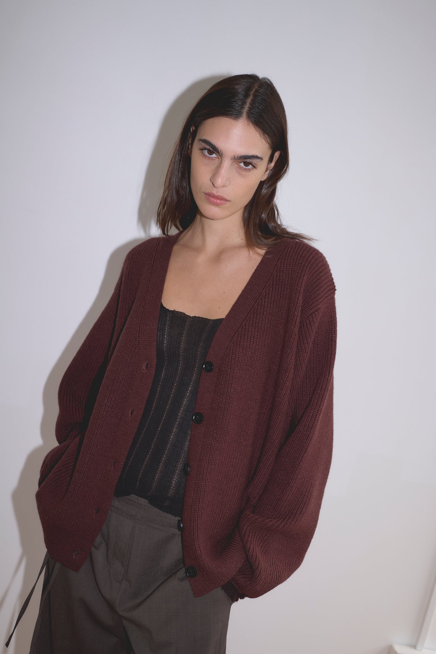 A woman with straight brown hair wears the Flounce Cardigan - Burgundy by Deiji Studios over a black top and brown pants, standing against a plain white background with a neutral expression.