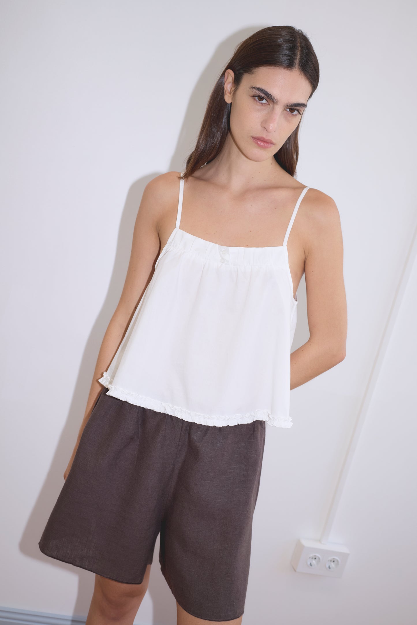 A model stands against a white wall wearing the Yoke Top - White with spaghetti straps and a ruffled hem, paired with dark brown linen bermuda shorts from Deiji Studios.