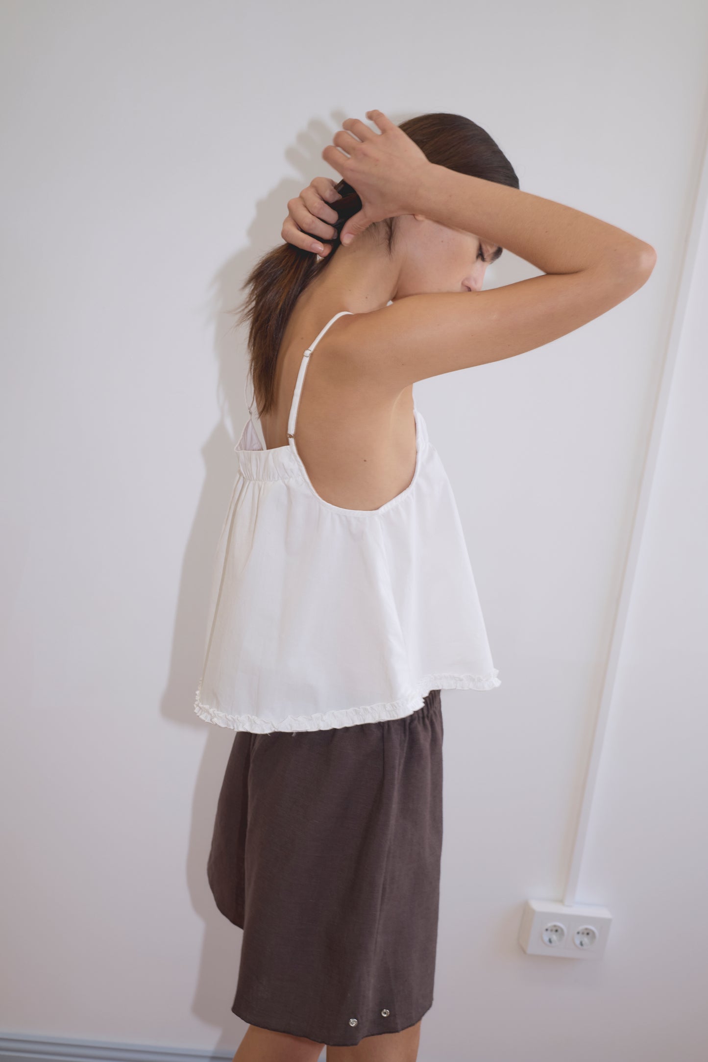 Wearing the Yoke Top - White, a person in dark shorts stands sideways against a white wall, lifting their hair with both hands. Their face is partially turned away, reflecting the relaxed style of Deiji Studios.