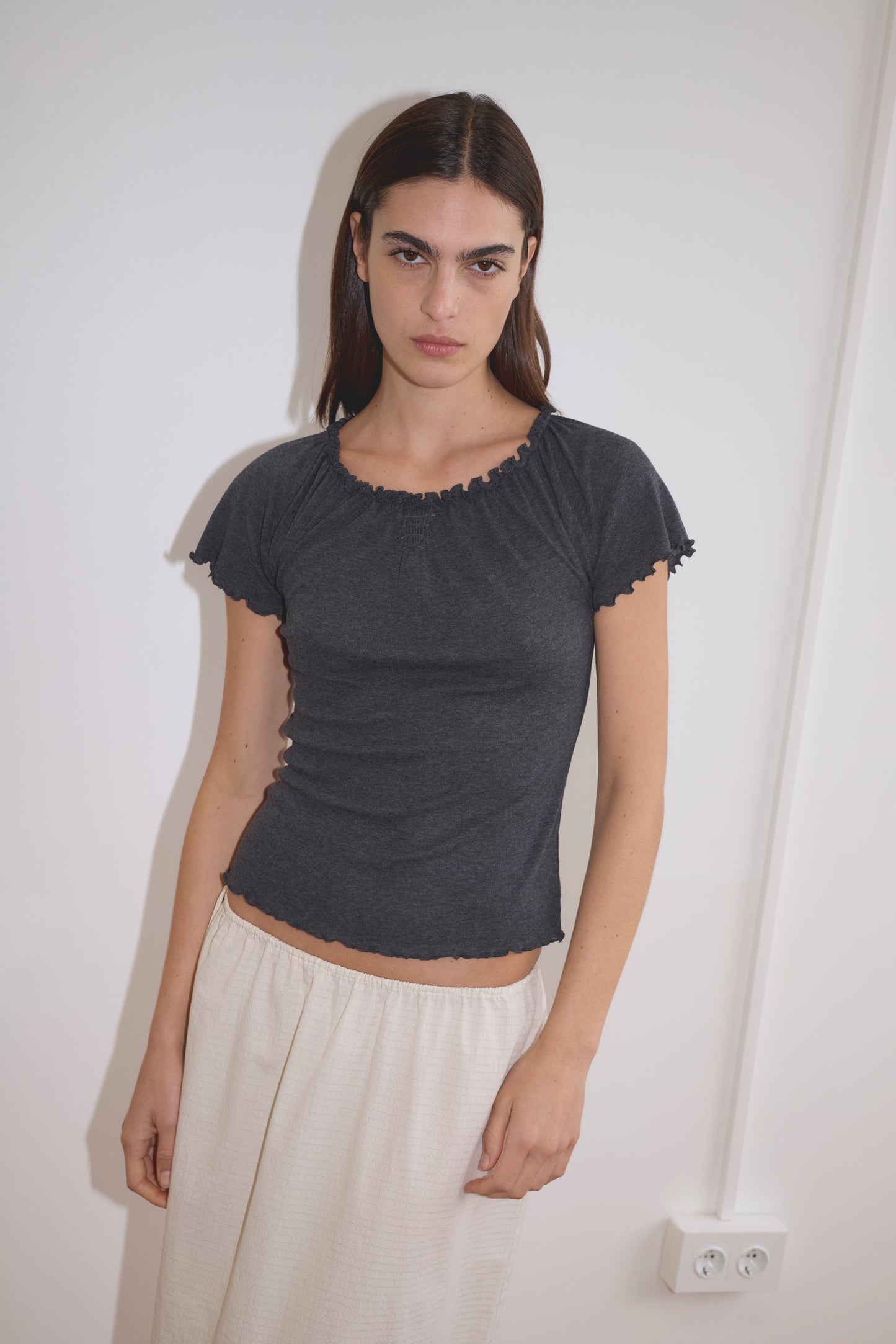 A woman with straight dark hair wears the Flounce Tee - Grey, paired with a light beige skirt, standing indoors against a white wall with an electrical outlet visible.