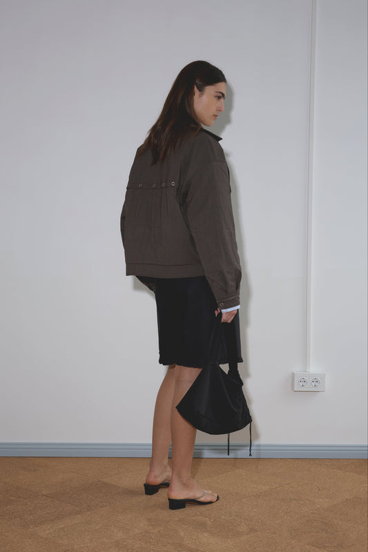 A woman stands indoors facing sideways, wearing the Lapel Jacket - Brown Stripe by Deiji Studios, a black skirt, and heeled sandals. She holds a black handbag on a beige floor against a plain white wall with an electrical outlet visible.