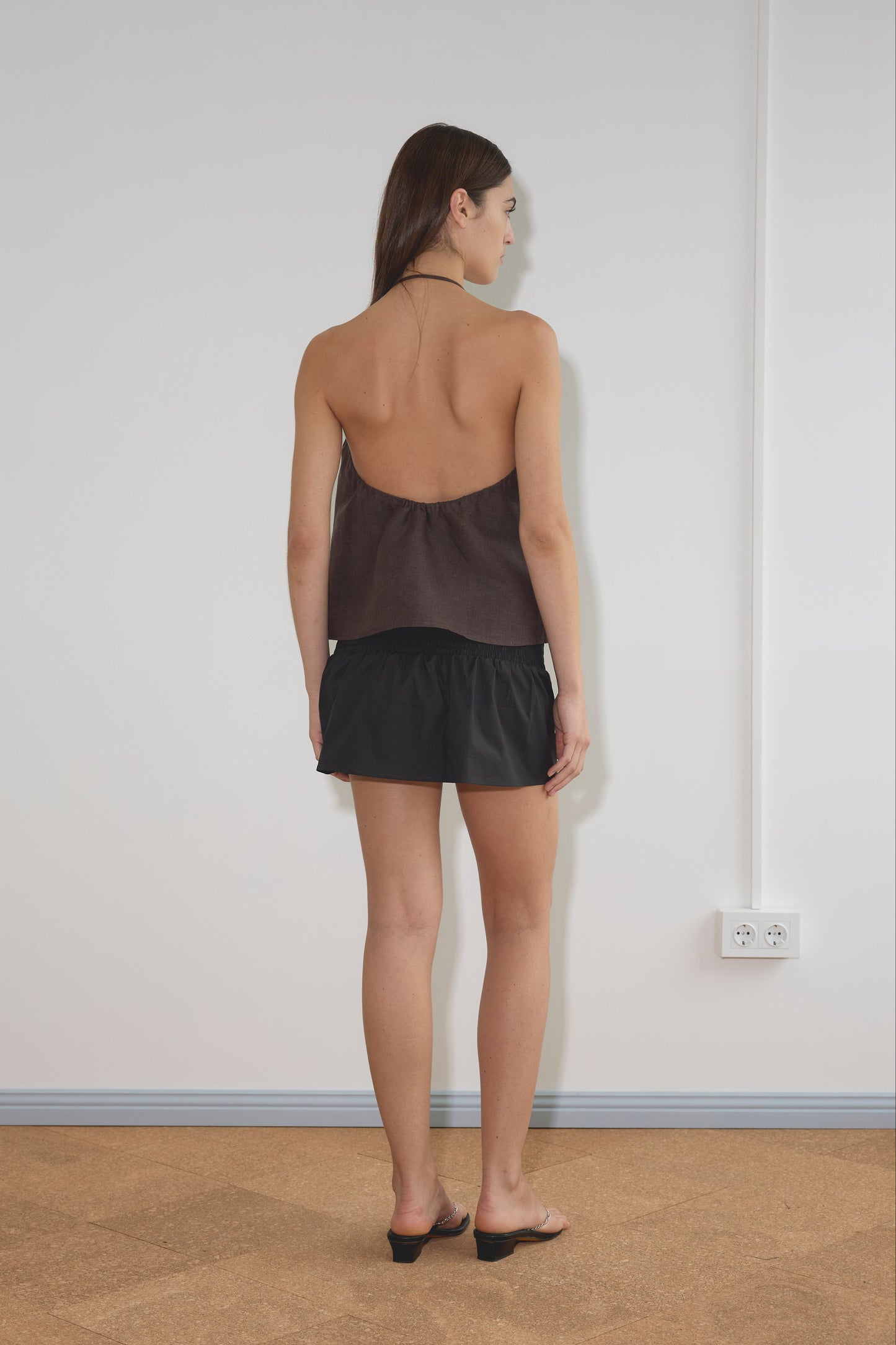 A woman with straight brown hair stands facing a white wall, wearing a dark halter top, the Shirring Skirt - Black, and black slide sandals in a light room with a power outlet and tan floor.