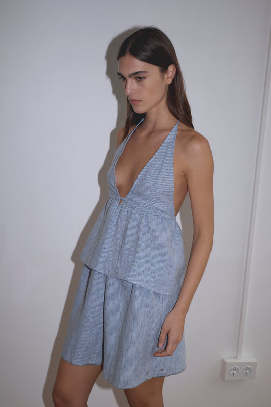 A woman with long dark hair wears the Linen Button Halter - Blue Stripe, a sleeveless, V-neck dress featuring subtle blue stripes, as she stands against a plain white wall.
