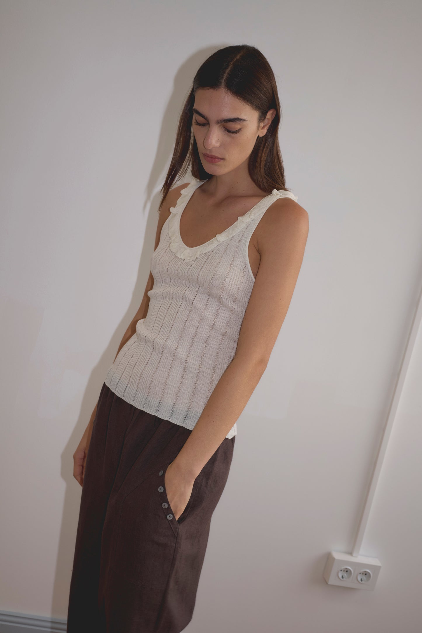 A woman with long dark hair, wearing the Deiji Studios Neck Ruffle Knit in Off White and loose dark pants, stands against a plain white wall with one hand in her pocket, looking down thoughtfully.
