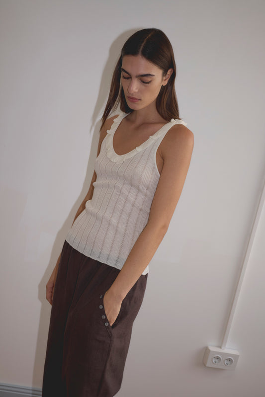Model wears the Neck Ruffle Knit - Off White by Deiji Studios, featuring a delicate neck ruffle, styled with a dark brown midi skirt with side buttons, standing against a white wall.