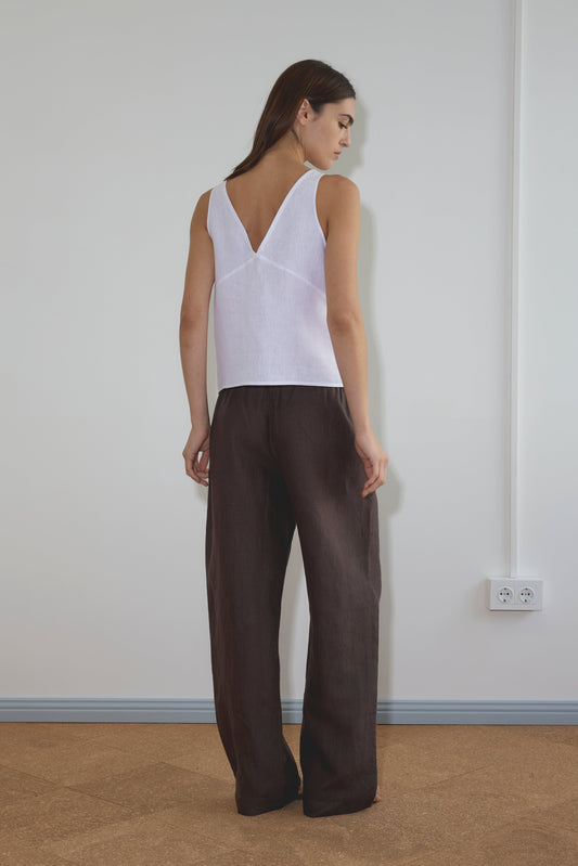 A woman stands facing away, wearing Deiji Studios' Two Part Top - White and loose dark brown pants in a minimal indoor space with white walls and a visible power outlet.