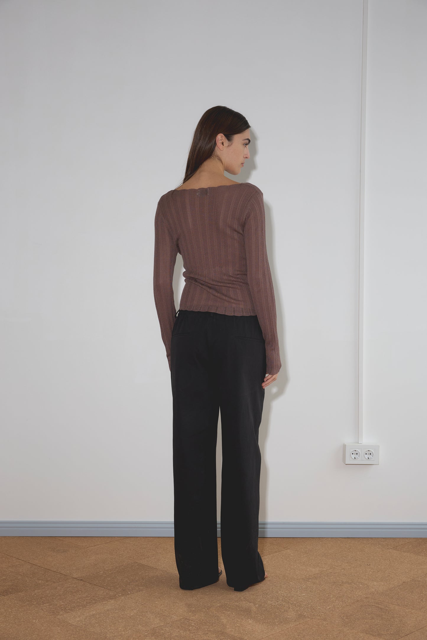A woman with long brown hair faces a white wall, wearing a sheer brown long-sleeve top and the Zip Pant - Black. An electrical outlet is visible on the wall near her right side.