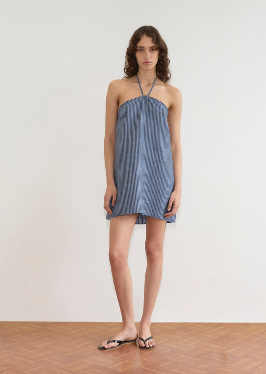 A person with wavy brown hair wears the Cross Over Dress, a short strapless blue and white striped design, paired with black sandals, standing indoors on a wooden floor against a plain white wall.