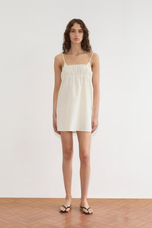 A person with wavy brown hair stands indoors on a wooden floor, wearing the sleeveless Edge Dress - Citrus Stripe and black sandals, facing forward against a plain white background.
