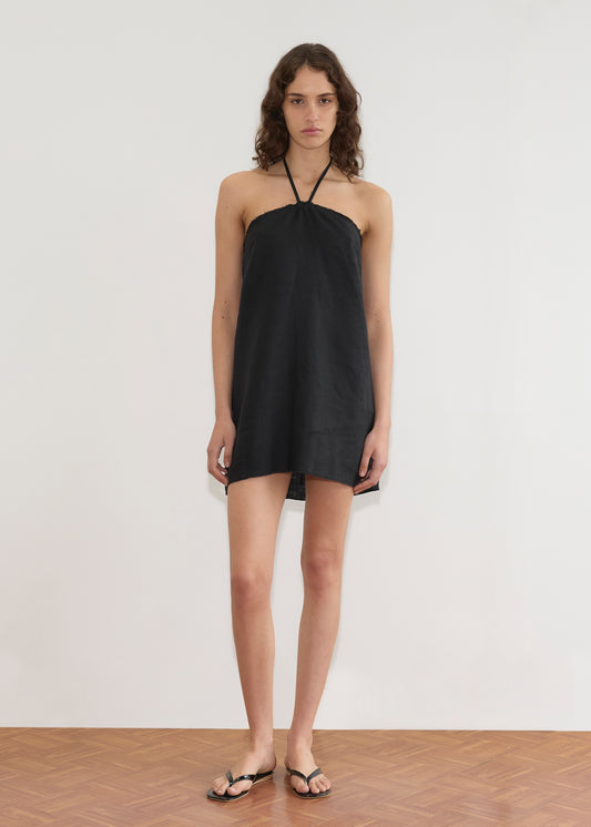 A person with wavy brown hair stands on a wood floor against a plain white wall, facing forward with a neutral expression. They wear the Cross Over Dress - Black from Deiji Studios and black sandals.