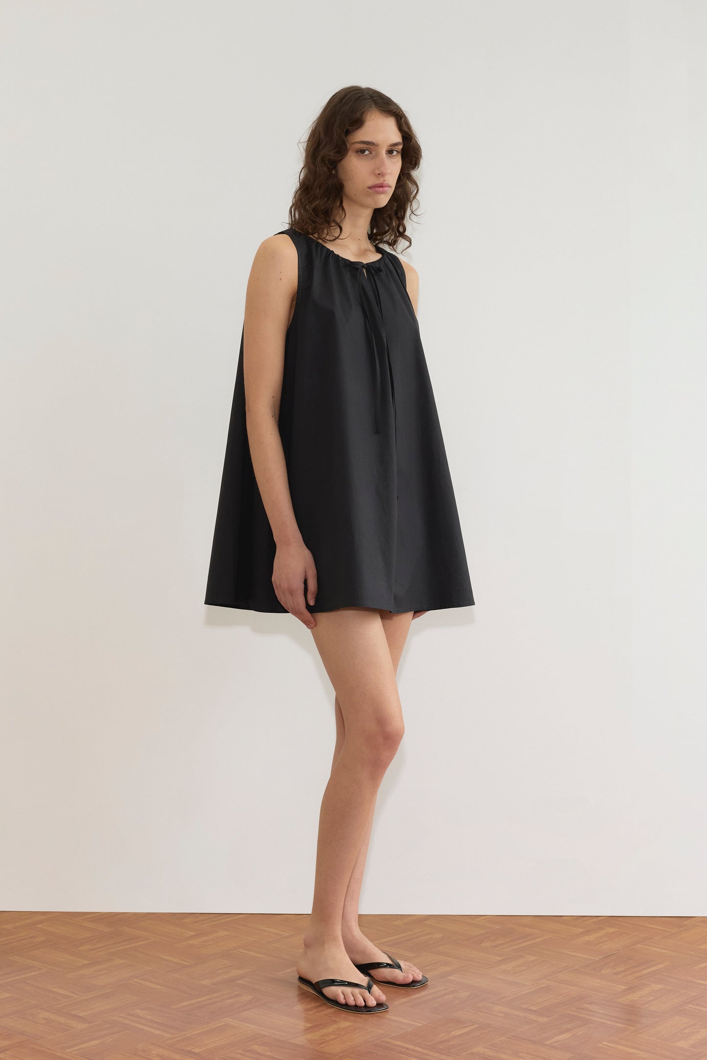 A woman with wavy brown hair stands indoors by a white wall, wearing the Deiji Studios Drawcord Dress - Black with an a-line silhouette and black flip-flops. She gazes to the side with a neutral expression.