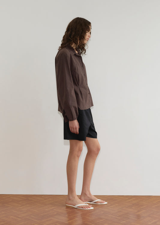 A person with long, wavy brown hair stands sideways against a plain white wall wearing the Cami Overshirt - Sepia over black shorts and white sandals, standing on a wooden floor.