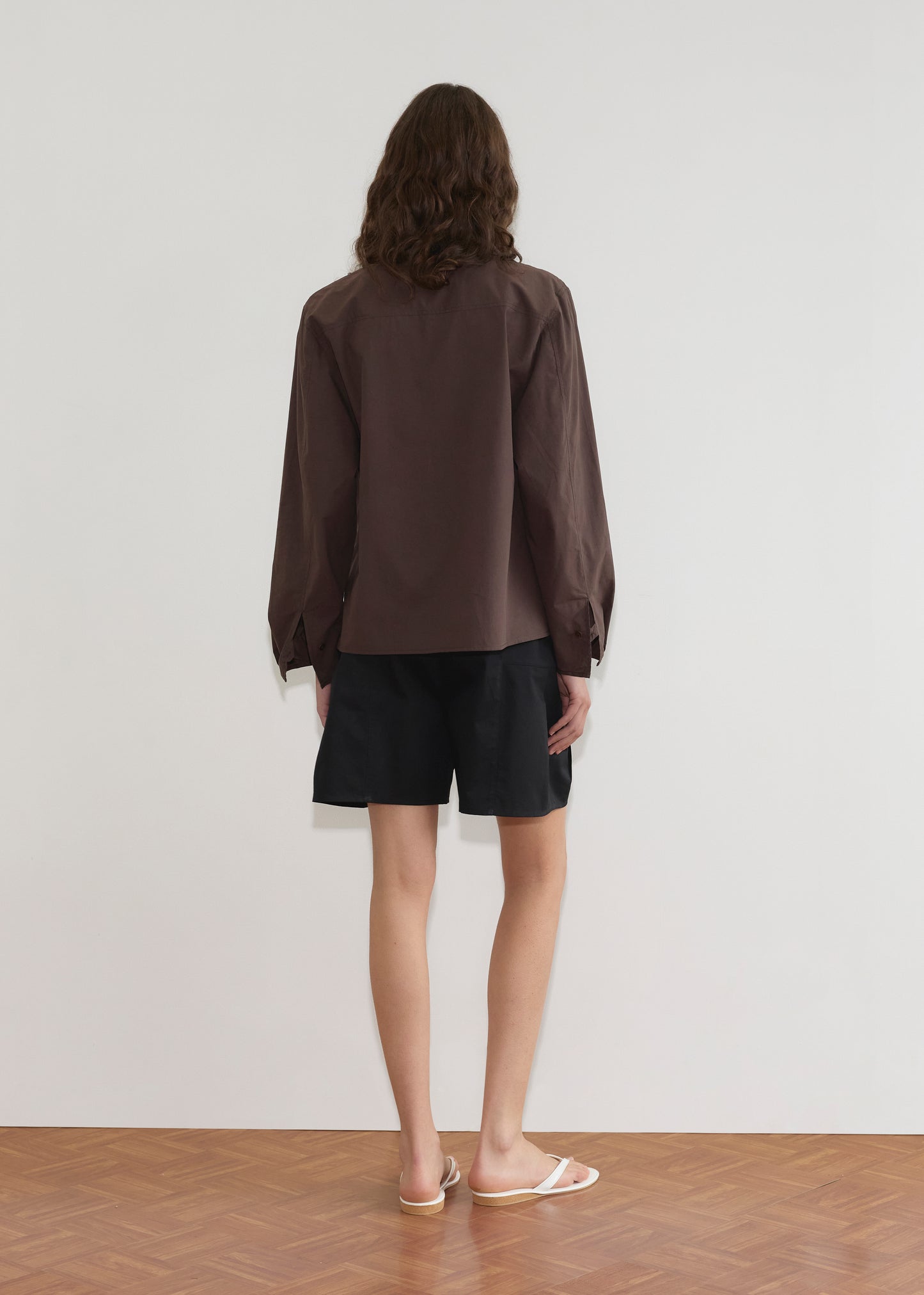A person with shoulder-length brown hair stands facing a white wall, wearing the Cami Overshirt - Sepia by Deiji Studios, black shorts, and white flip-flops on a wooden floor.