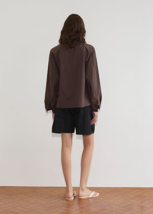 A person with shoulder-length brown hair stands facing a white wall, wearing the Cami Overshirt - Sepia by Deiji Studios, black shorts, and white flip-flops on a wooden floor.
