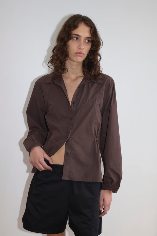 A person with wavy brown hair stands against a white wall, wearing the Deiji Studios Cami Overshirt in Sepia partially open over black shorts, looking slightly to the side with a neutral expression.