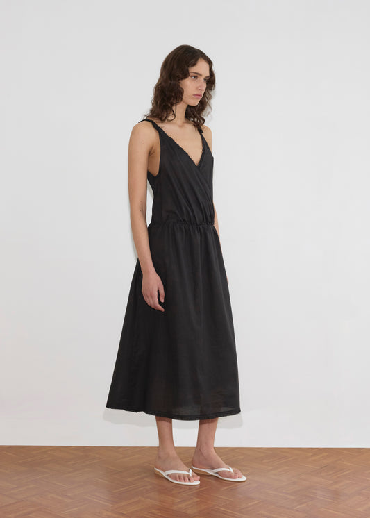 A person with wavy brown hair wears the Crossed Dress - Black from Deiji Studios, a sleeveless midi dress with a crossover v-neckline, paired with white sandals, standing on a wooden floor against a plain white background.