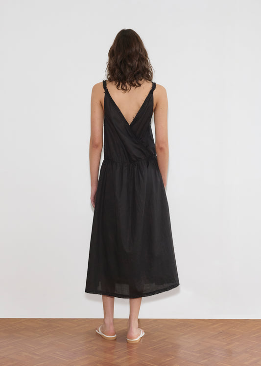 A woman with shoulder-length wavy hair stands facing a white wall, wearing the Crossed Dress - Black by Deiji Studios with a crossover v-neckline and V-back, paired with light-colored flat sandals on a wooden floor.