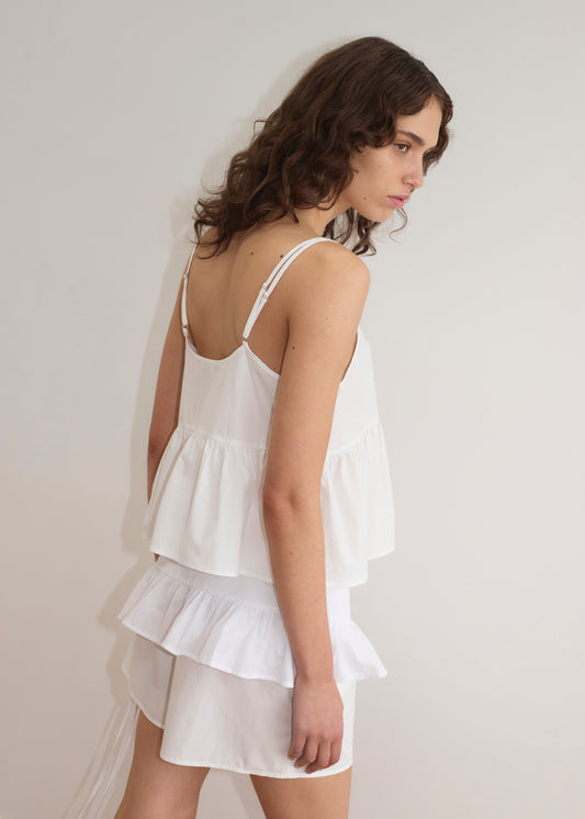 A woman with wavy brown hair stands sideways against a plain white wall, wearing the Flounce Belt - White as a sleeveless ruffled top with adjustable ties and paired with a matching skirt.