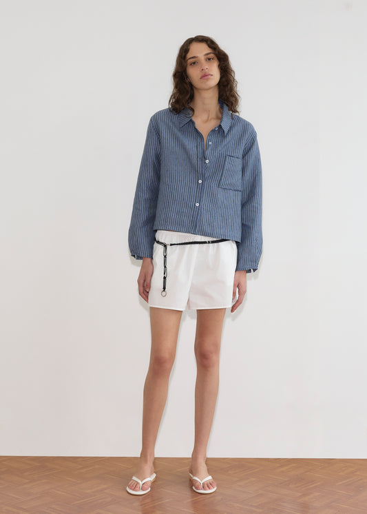 A woman with wavy brown hair and a relaxed expression stands against a plain white wall wearing the Double Placket Shirt - Azure Stripe, paired with white shorts, a black belt, and white sandals.