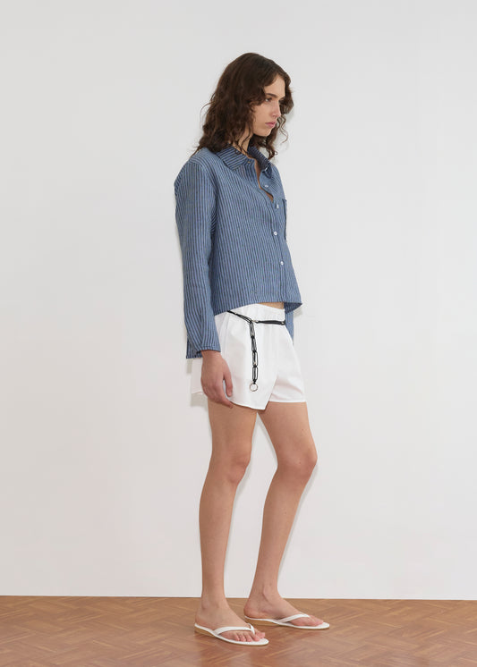 A person with wavy brown hair wears the Double Placket Shirt - Azure Stripe, paired with white shorts featuring a black chain and white flip-flops, standing indoors on a wooden floor against a plain white wall.