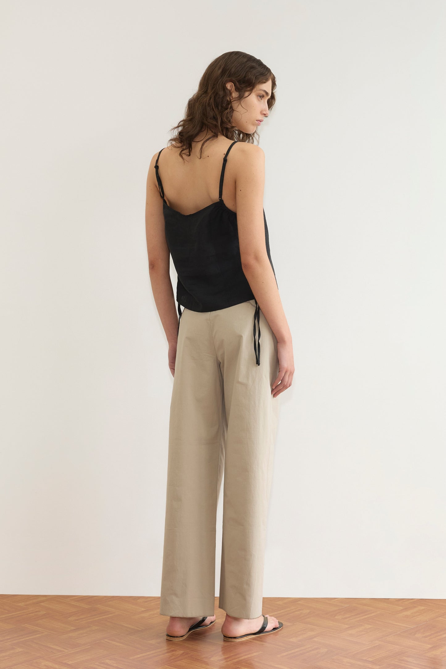 A woman with wavy brown hair stands facing away, wearing the V Neck Cami - Black from Deiji Studios, beige wide-leg pants, and black flip-flops on a wooden floor with a plain white wall backdrop.
