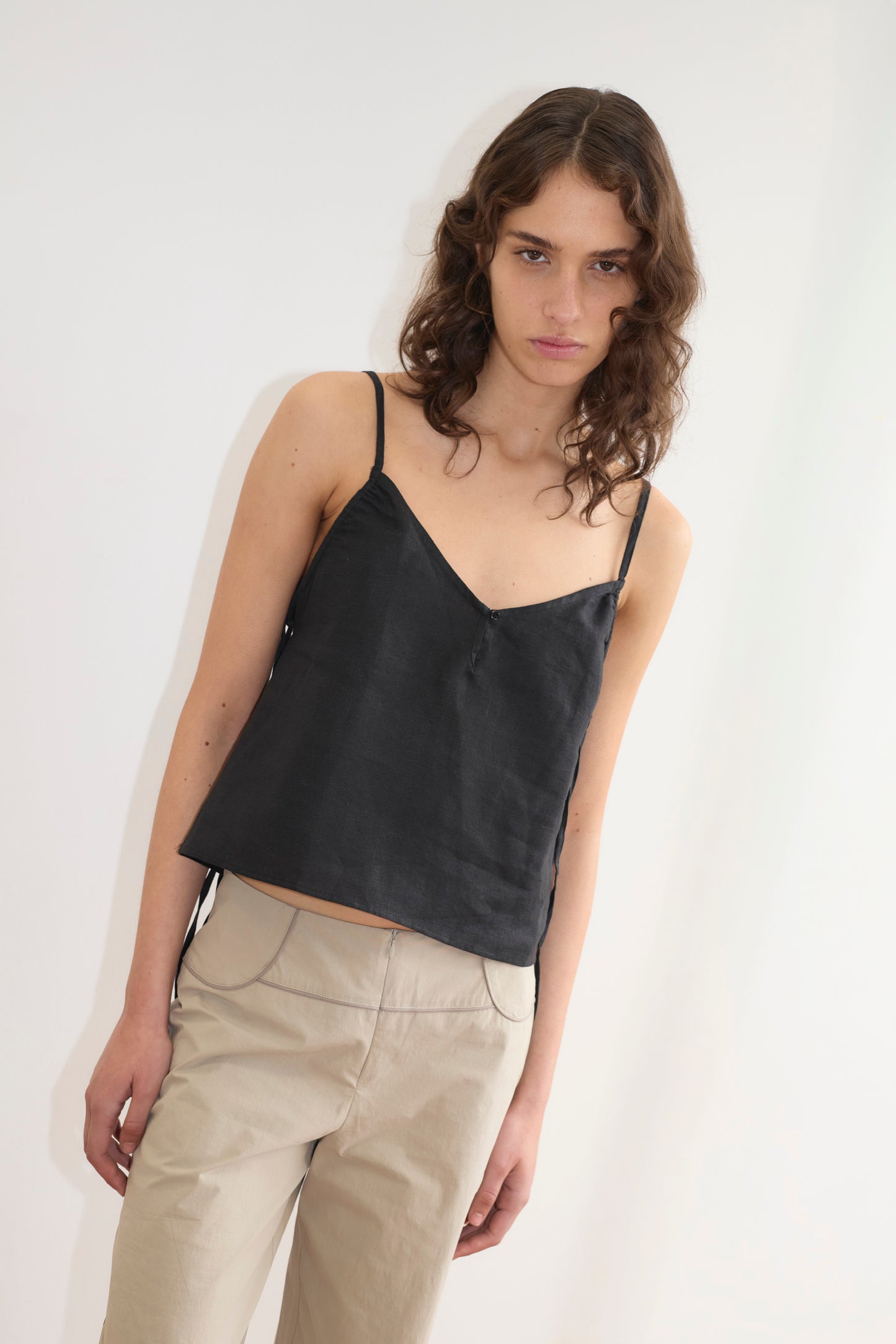 A model with curly brown hair wears the V Neck Cami in black satin with spaghetti straps and a V-neckline, paired with beige wide-leg trousers, against a white background.