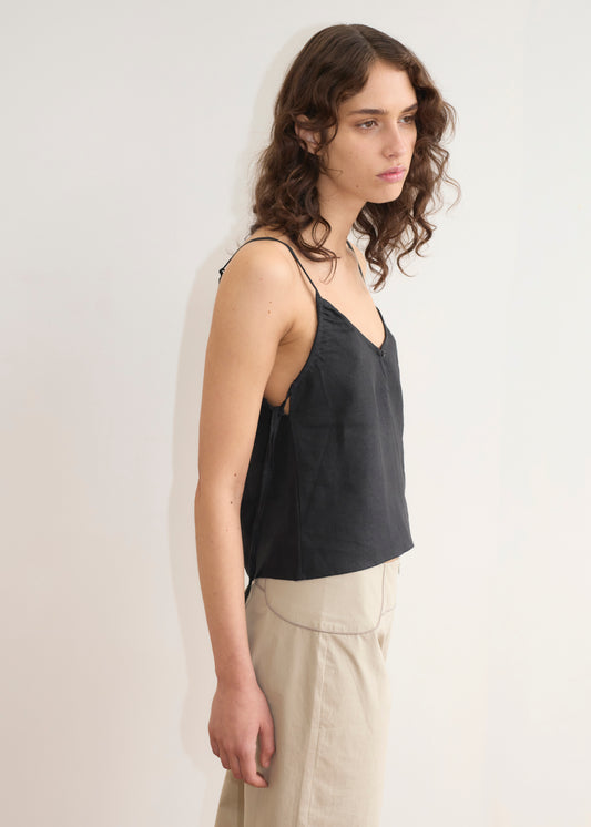 A woman with long, wavy brown hair stands in profile against a plain light background, wearing the V Neck Cami - Black from Deiji Studios and high-waisted beige pants.