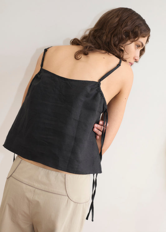 A person with wavy brown hair, wearing Deiji Studios' V Neck Cami - Black and beige pants, stands against a plain light-colored wall, facing away and looking over their shoulder.