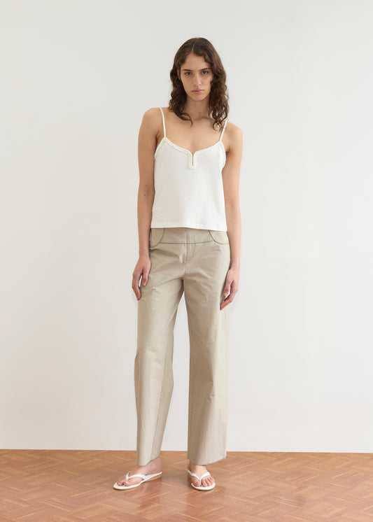 A woman stands indoors on a wooden floor, wearing a white camisole top and the Deiji Studios Panelled Pant - Stone with a straight-leg silhouette. She has long wavy hair and a neutral expression, set against a plain light background.