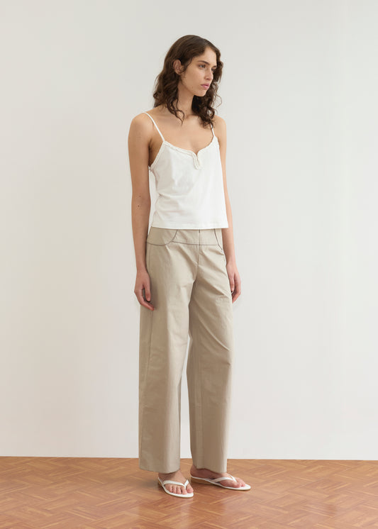 A woman stands indoors in a white camisole top, Deiji Studios’ Panelled Pant - Stone with a straight-leg silhouette, and white sandals. She has wavy brown hair and looks to the side against a plain light background.