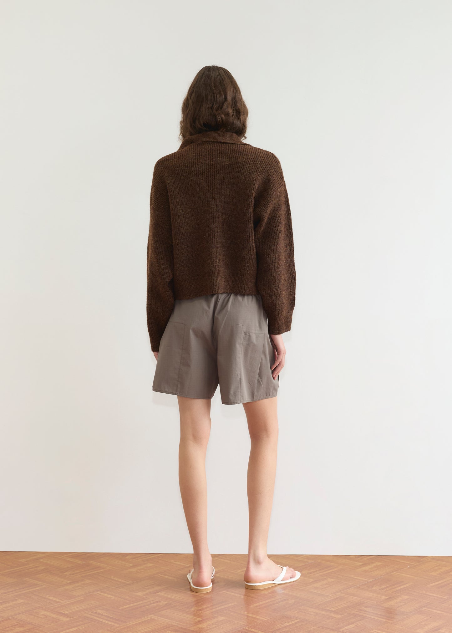 A person with wavy brown hair stands facing away, wearing a Deiji Studios Double Layer Cardigan in brown, loose gray shorts, and white flip-flops against a plain white wall and wooden floor.