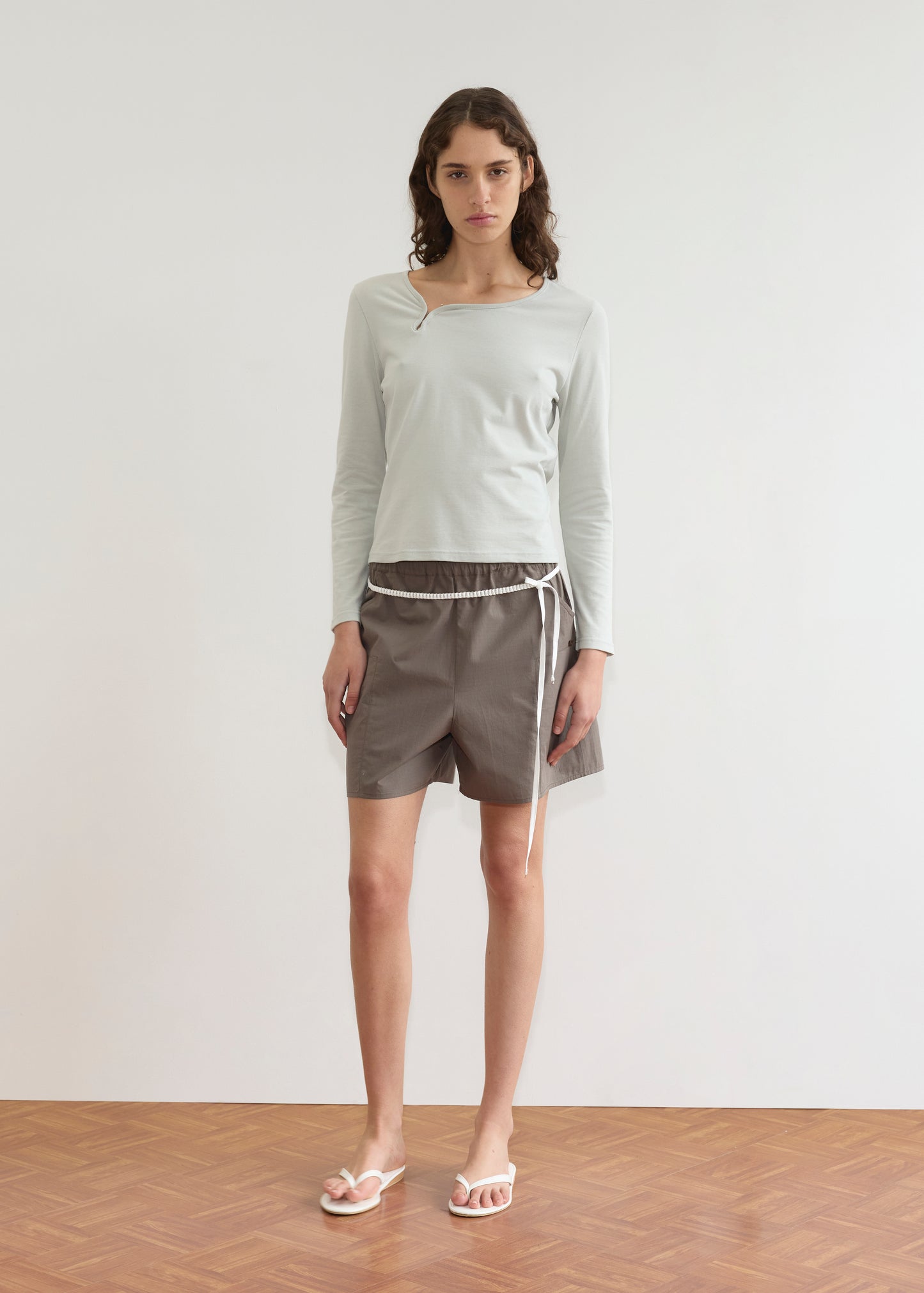 A person with long, wavy brown hair stands indoors against a plain white wall, wearing the Clasp Longsleeve - Grey Blue by Deiji Studios, gray drawstring shorts, and white flip-flops on a wooden floor.