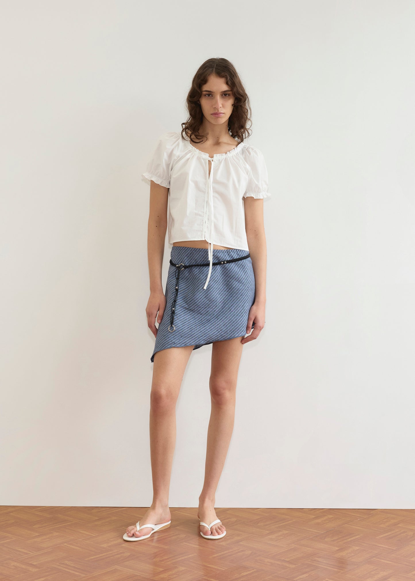 Indoors, a person stands against a plain white wall on a wooden floor, wearing the Close Shirt in White, paired with a blue patterned mini skirt featuring an asymmetrical hem and white sandals.
