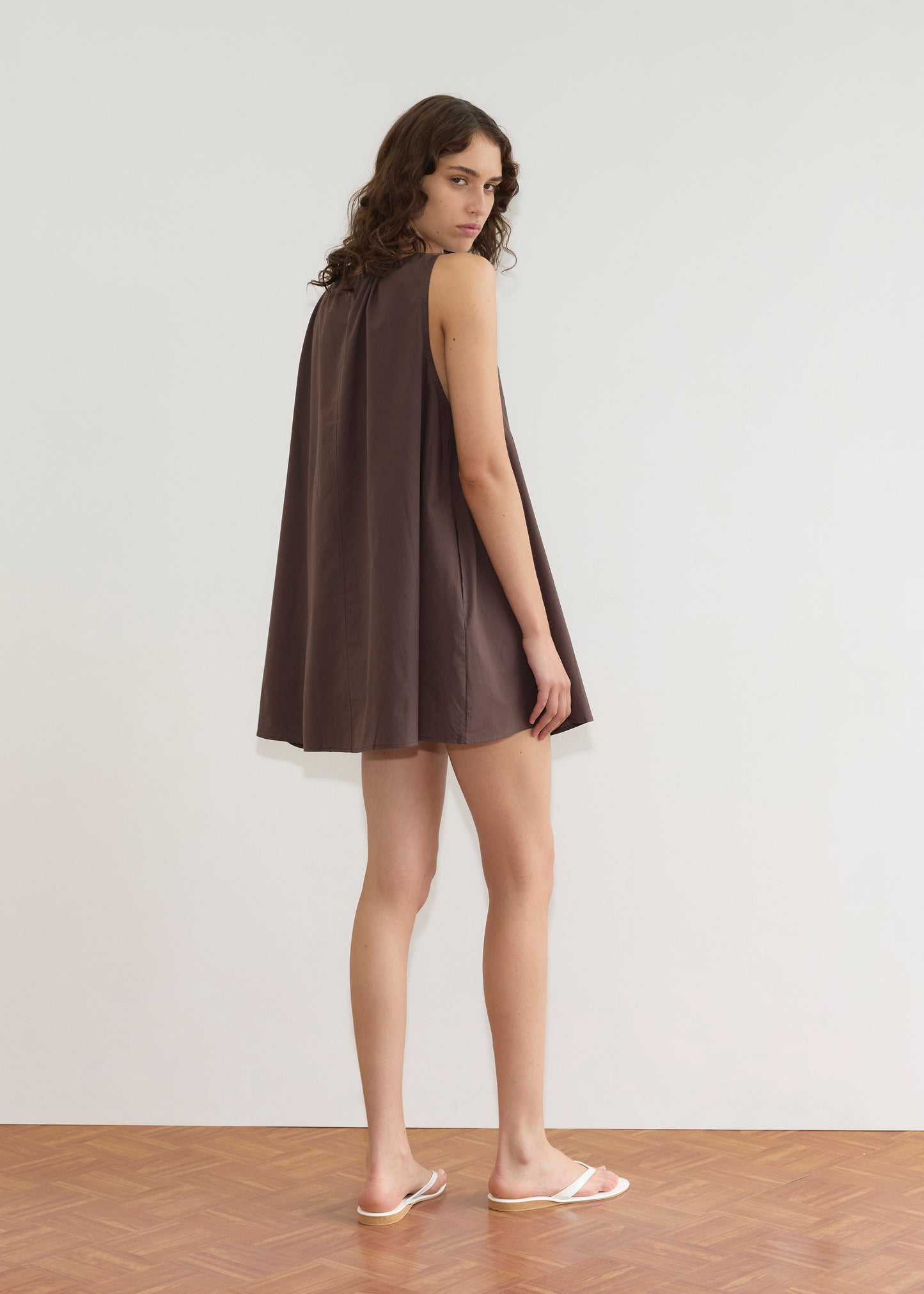 A person with long, wavy brown hair stands sideways on a wooden floor, wearing the Deiji Studios Drawcord Dress - Sepia with an a-line silhouette and white flip-flops, posed against a plain white wall.