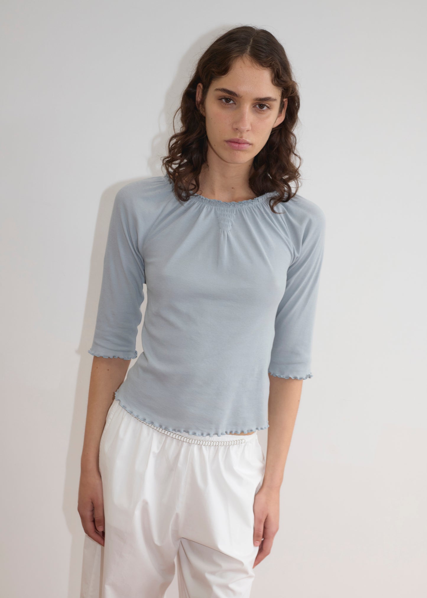 A person with wavy brown hair wears Deiji Studios’ Cotton Flounce 3/4 Sleeve - Baby Blue top and loose white pants, standing against a plain white background with a neutral expression.