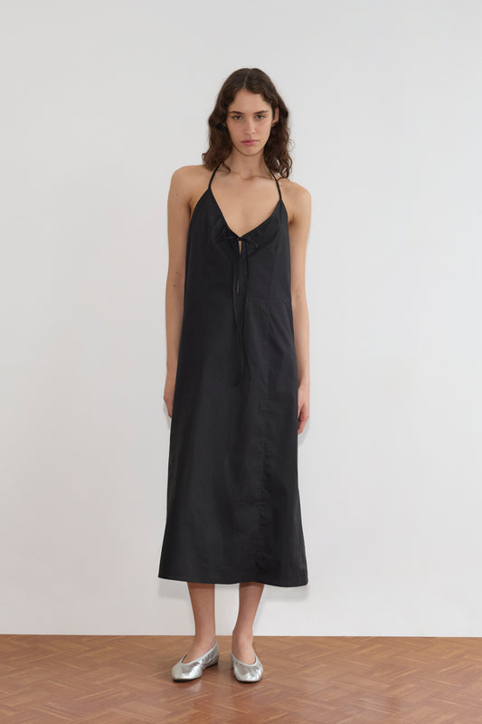 A woman with long brown hair wears the Seamed Halter Dress - Black, styled with silver ballet flats. She stands indoors on a wooden floor against a plain white wall.