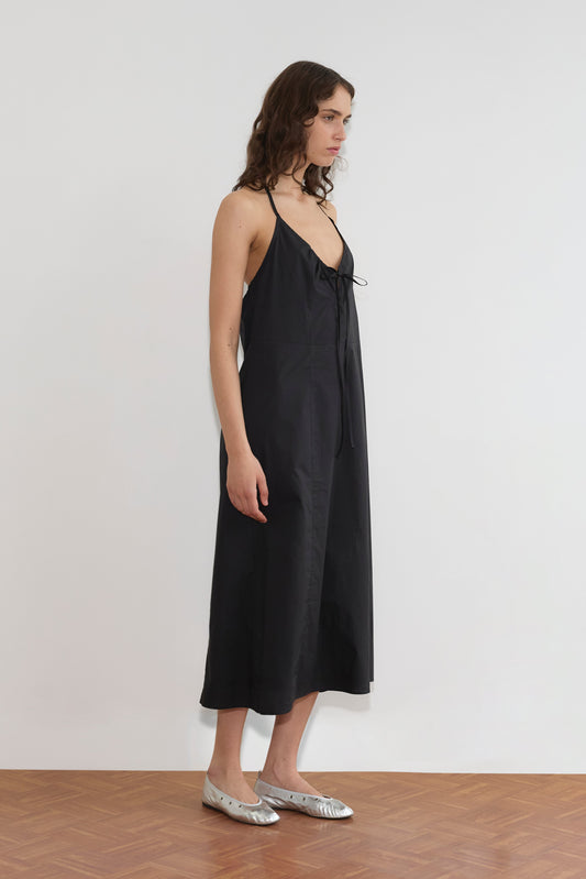 A woman stands sideways against a plain white wall, wearing the Seamed Halter Dress - Black with thin straps and bow detail, paired with silver flat shoes on a wooden floor.