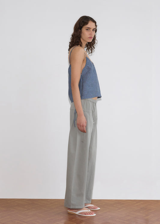 A person with wavy brown hair stands sideways in a Deiji Studios V Neck Cami - Azure Stripe, paired with loose light gray pants and white sandals, set against a plain white wall and wooden floor.