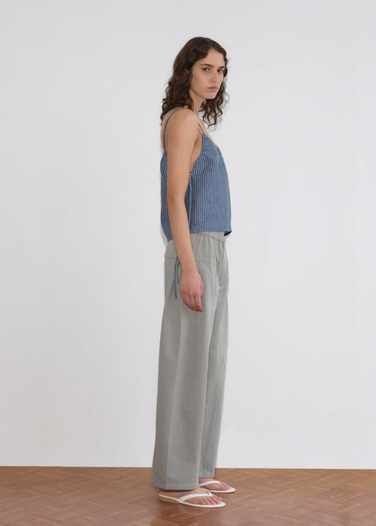 A person with wavy brown hair wears a blue striped sleeveless top, the Pleat Pant - Sage Grey, and white sandals, standing sideways against a plain white background on a wooden floor.