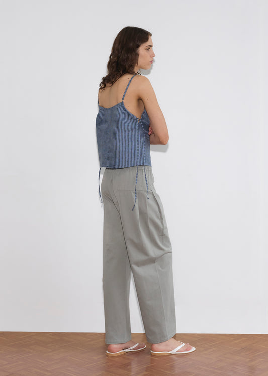 A person with long brown hair stands sideways against a white wall, wearing the V Neck Cami - Azure Stripe by Deiji Studios, light gray high-waisted pants, and white sandals on a wooden floor.