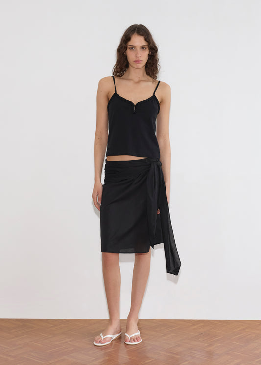 A woman stands indoors against a plain white wall, wearing the Clasp Singlet - Black with adjustable straps, a black wrap skirt tied at the side, and white flip-flops. Her wavy hair and the wooden floor complete the look.