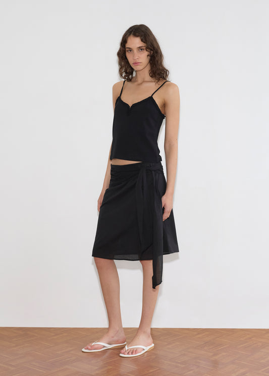 A woman with wavy brown hair wears a black camisole top, the Deiji Studios Scarf Midi - Black skirt tied at the waist, and white flip-flops. She stands on a wooden floor against a plain white wall.