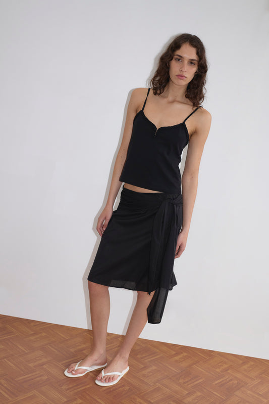 A woman stands indoors on a wooden floor, wearing the Clasp Singlet - Black by Deiji Studios with adjustable straps, a black wrap skirt, and white flip-flops. She has wavy brown hair and a neutral expression, one hand resting by her side.