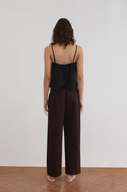 A person with wavy brown hair stands facing a white wall, wearing a black sleeveless top, Pocket Jean - Dark Brown by Deiji Studios, and white heeled sandals on a wooden floor.