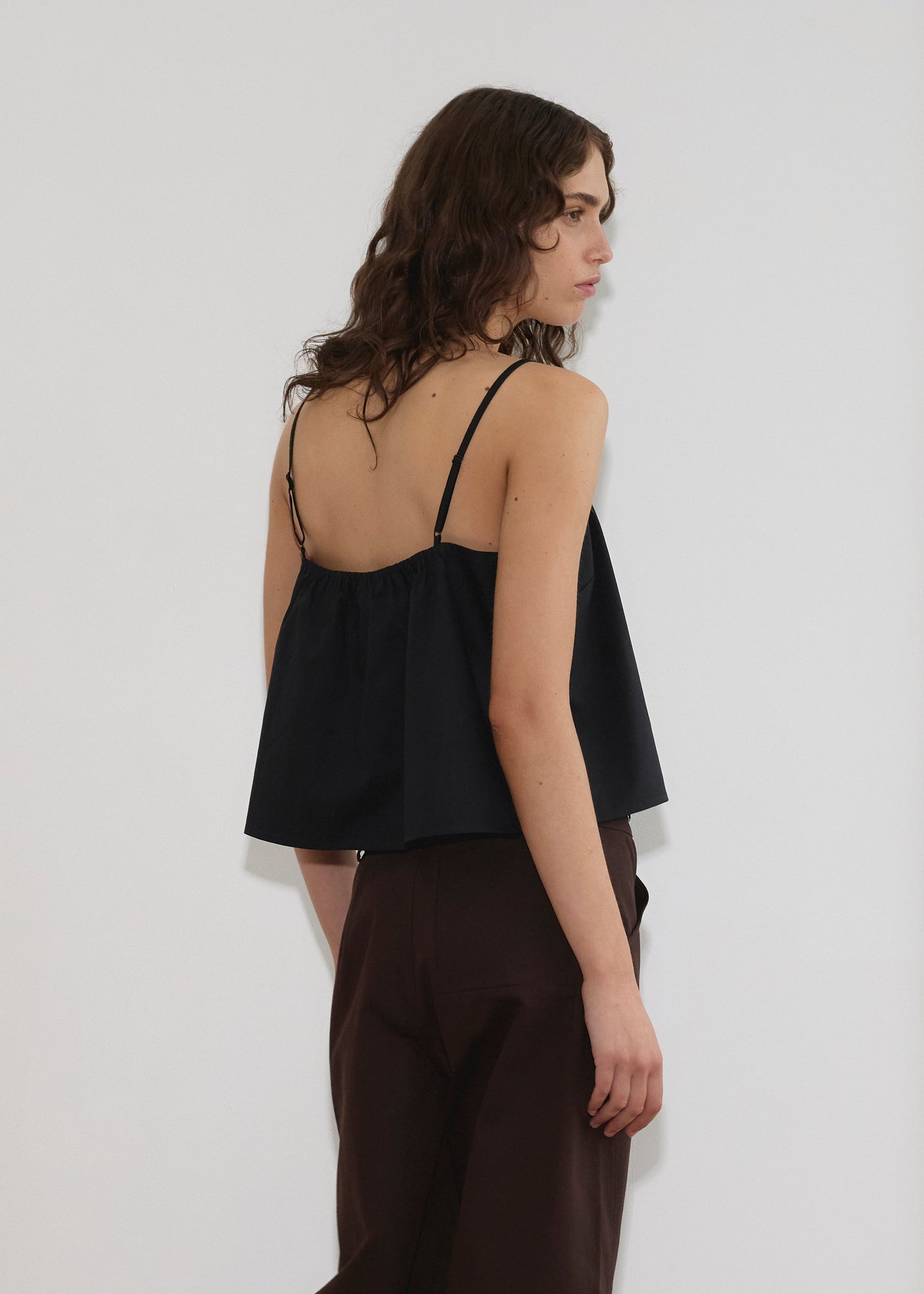 A person with wavy brown hair stands sideways, wearing the Double Layer Top - Black by Deiji Studios, wide-legged dark brown pants, and white sandals in a minimalist room with wooden floors and white walls.