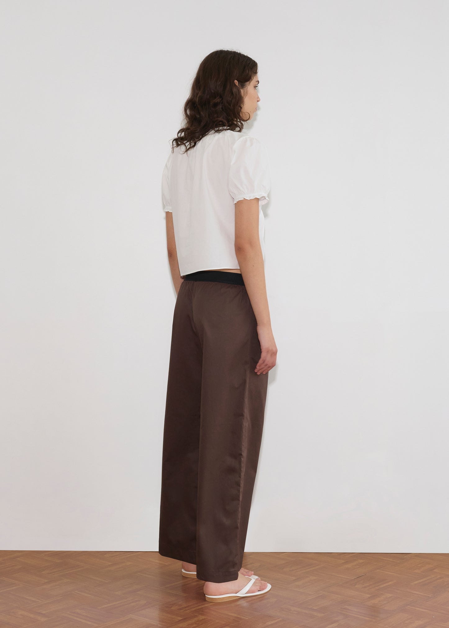 A woman with wavy brown hair stands on a wooden floor, facing away. She wears a white blouse and the Contrast Waist Pant - Brown from Deiji Studios, paired with white sandals against a plain white wall background.