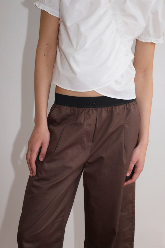 A person wearing a white short-sleeved blouse and the Contrast Waist Pant - Brown by Deiji Studios stands against a plain background, showing only their torso and upper legs.